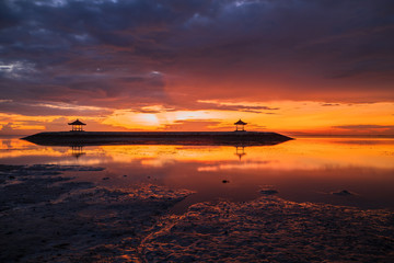 Sunrise seascape. Traditional gazebos on an artificial island in the ocean. Water reflection. Bright sunlight at horizon. Couldy sky. Sanur beach, Bali, Indonesia.
