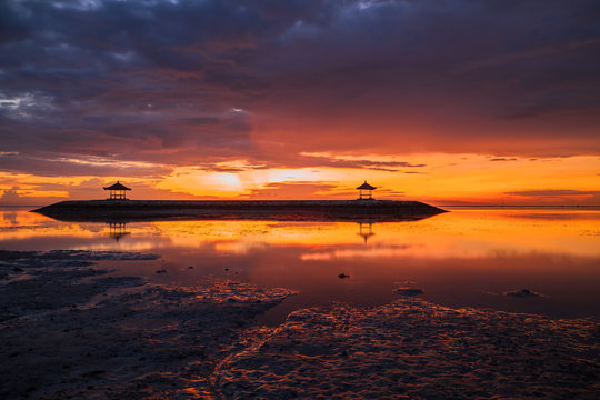 Sunrise seascape. Traditional gazebos on an artificial island in the ocean. Water reflection. Bright sunlight at horizon. Couldy sky. Sanur beach, Bali, Indonesia.