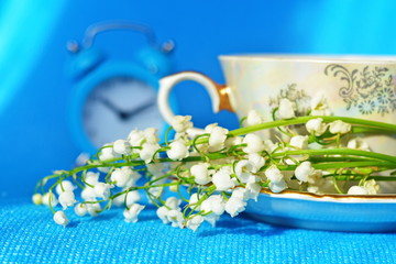 Bunch white flower of lily of the valley on saucer next teacup on blue background with blurred...