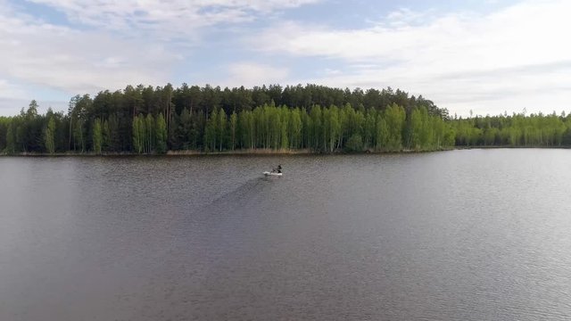 03. Aerial view of lake surrounded by a forest. Two fishermens in a rubber inflatable boat fishing. One fisherman rowing oars, the other stands with a fishing rod. A sunny summer day
