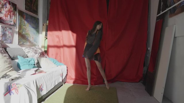 young barefoot ballerina in short clothes dances near red fabric backdrop in sunny art studio with drawings slow motion