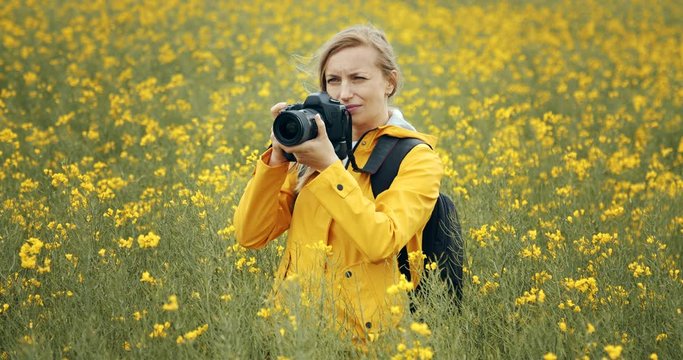 Front view of smiling mature lady in yellow jacket with big backpack using digital camera for taking pictures of green field. Concept of photography and working process