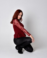 Portrait of a pretty girl with red hair wearing leather pants and long sleeved lace shirt.  full length kneeling pose  isolated against a studio background