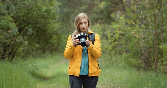 Female photographer with blond hair standing among wild green forest and taking pictures of beautiful nature around. Mature lady using digital camera for shooting outdoors.