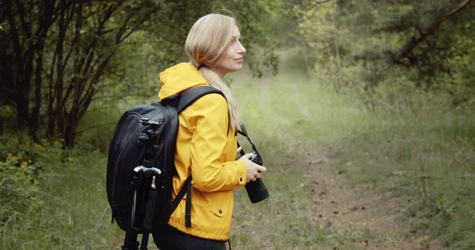 Smiling mature woman with big backpack taking photos of wild nature at spring forest. Female photographer with blond hair walking among green trees and using digital camera.