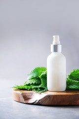 Natural spa, herbal cosmetics. Nettle lotion, cream, shampoo or soap in white bottle and fresh nettles leaves on grey background. Medicinal herb for health and beauty, skin care and hair treatment.
