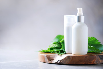Natural spa, herbal cosmetics. Nettle lotion, cream, shampoo or soap in white bottle and fresh nettles leaves on grey background. Medicinal herb for health and beauty, skin care and hair treatment.
