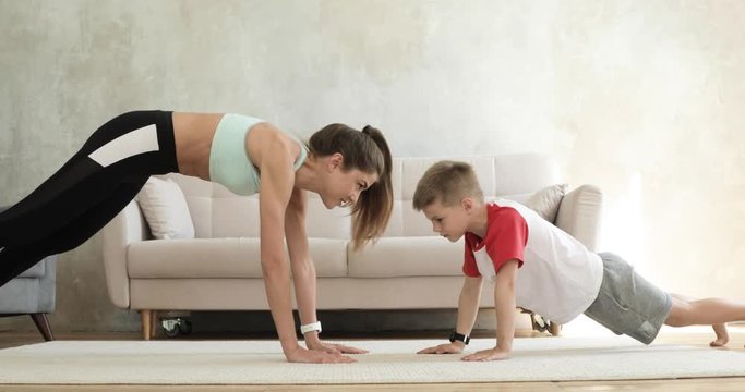 Training of mom and son making plank exercise at home giving five each other, side view. Sporty fit family doing fitness aerobic exercises in living room. Home workout, training and wellness concept.