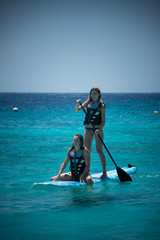 2 girls on a paddle board while wearing bikinis and lifejackets. Blue open ocean and blue sky.