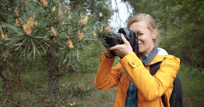 Cheerful mature lady with blond hair taking photos of pine trees while standing among green forest. Female photographer in yellow jacket travelling with backpack and professional equipment