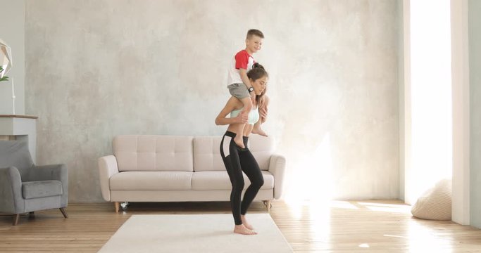 Young sporty woman mother is making lunges with her son boy sitting on her shoulders. Home workout, training with children and wellness concept. Motherhood and self-development.