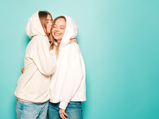 Two young beautiful blond smiling hipster girls in trendy summer hoodie clothes. Sexy carefree women posing near blue wall.Model kissing her friend in head