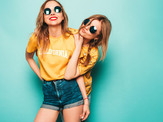 Two young beautiful smiling hipster girls in trendy summer yellow T-shirts. Sexy carefree women posing near blue wall. Trendy and positive models having fun in sunglasses