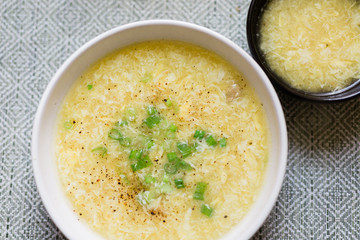 A large bowl of egg drop soup topped with green scallions. A smaller bowl is in the corner. 