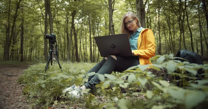Happy mature lady in casual clothing sitting among green forest and using personal laptop. Professional photographer with blond hair enjoying working time outdoors.