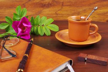 Morning coffee. Cup of coffee, notebook, pen, glasses, flash drive, rosehip flower on a wooden table.