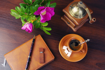 Cup of coffee, notebook, pen, flash drive, coffee grinder, rose hip flower on a wooden table.