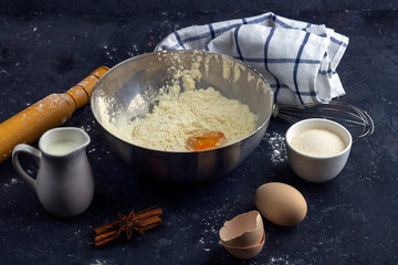 Flour with egg in a metal bowl among ingredients and utensils for cooking cake (flour, egg, milk, sugar, rolling pin, towel) on dark table. The concept of making dough for baking. Close up