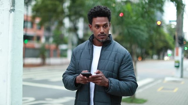 Black guy looking at smartphone device walking in sidewalk, mixed race person messaging and walking