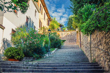 Fototapeta na wymiar Cobblestone staircase with stairs, green trees, bushes and flowers, street lights between stone walls in Brescia city historical centre, Lombardy, Northern Italy