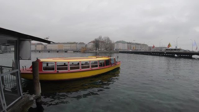 Genevan yellow boat named mouettes, public transportation leaving the port in the harbor of Geneva, under a gray sky, with a seagull on the roof.