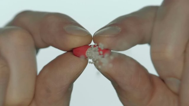 A man opens a red medical capsule and spills white powder from it directly into the camera on a white background. Closeup. Slow motion. High speed camera