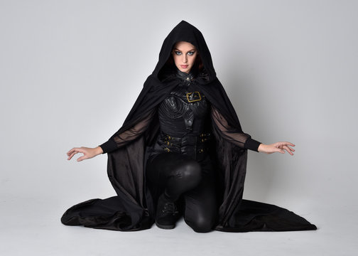 Fantasy Portrait Of A Woman With Red Hair Wearing Dark Leather Assassin Costume With Long Black Cloak.  Full Length Kneeling Pose, Isolated Against A Studio Background.