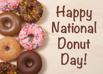 Flat lay top view various flavors cake donuts frosted, laying on light wood table with  HAPPY NATIONAL DONUT DAY text