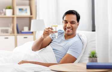 people, bedtime and rest concept - happy smiling indian man lying in bed at home and taking glass of water with lemon from table