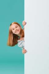 a laughing girl standing against a turquoise background holds an empty white Board in her hand and looks out from behind it