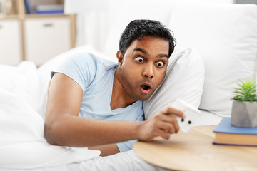 people, bedtime and rest concept - overslept indian man looking at alarm clock lying in bed at home