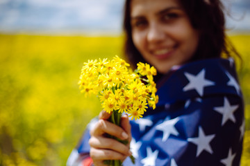 Patriotic young woman wrapped in an american flag with a bouquet of yellow flowers on a beautiful field. USA independence day 4th of July celebration. Summer holidays concept.
