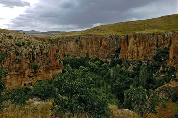 Ihlara Canyon in Cappadocia. The grass-covered plain is divided into parts by a gorge with steep rocky slopes. At the bottom, trees grow. Cloudy. In the distance are the silhouettes of mountains.
