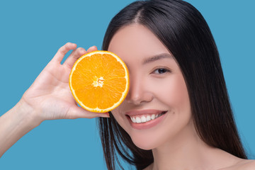 Smiling cute young woman holding a slice of orange and feeling pleased