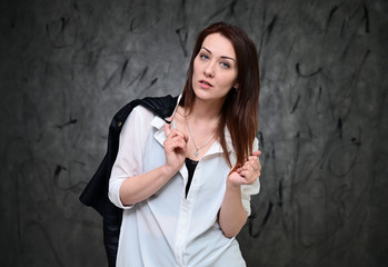 Fototapeta premium A pretty model is standing on a gray background in a white shirt holding a black jacket. Studio portrait of a caucasian brunette girl with minimal makeup.