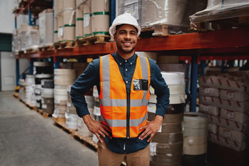 Portrait of happy manager standing with his hands on waist wearing white helmet and safety vest in...