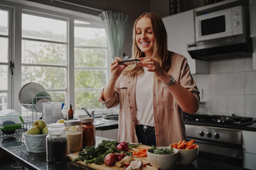 Young woman taking photo of her food with smart phone in modern kitchen at home during isolation...