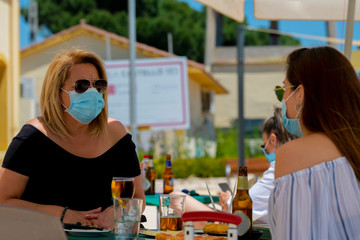 Women using surgical masks and sunglasses sitting at an outdoor bar terrace with beers and snacks