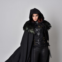 Close up fantasy portrait of a woman with red hair wearing dark leather assassin costume with long black cloak.  isolated against a studio background.