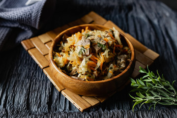Stewed cabbage with oyster mushrooms and pork tongue with carrots and onions. Hot dish in a wooden plate on a wooden stand, next to a fork, a sprig of grass and a napkin. Black background with relief