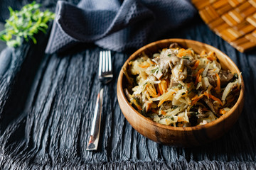 Stewed cabbage with oyster mushrooms and pork tongue with carrots and onions. Hot dish in a wooden plate on a wooden stand, next to a fork, a sprig of grass and a napkin. Black background with relief