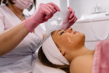 Obraz na płótnie Canvas Calm peaceful woman lying during beauty spa procedure. Hands in white gloves put some white mask all over face for beauty procedure and skin treatment