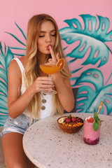 Caucasian girl spending time in cafe, drinking orange cocktail, touch her lips with fonger, sitting by the table with tropical smoothie bowl. Lifestyle portrait of a beautiful, young and attractive 