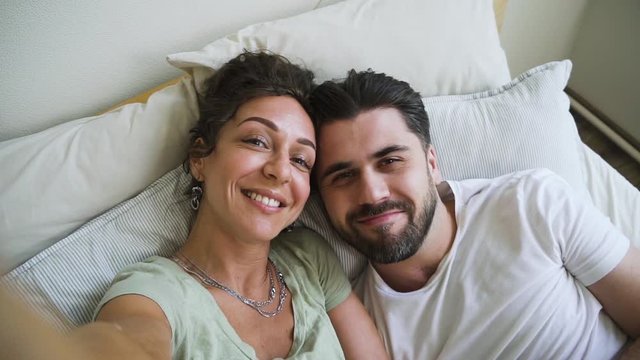 Happy american couple take selfie and kiss while lying on bed in white bedroom spbd. Bearded man and attractive woman are having fun together, kissing and taking photo, spending time in home interior