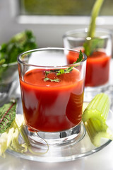 Two glasses of tomato juice with parsley and celery decorations, stand near the window, morning sunlight shines, shallow depth of field, selective focus. Natural drinks concept.
