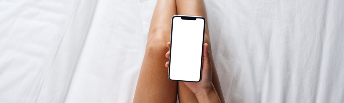 Top view mockup image of a woman holding white mobile phone with blank desktop screen while lying on a cozy white bed at home