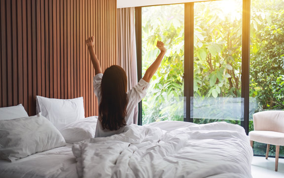 Rear view image of a woman do stretching after waking up in the morning  , looking at a beautiful nature view outside bedroom window