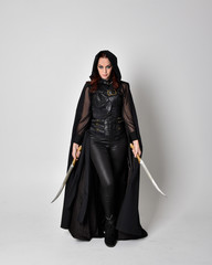 fantasy portrait of a woman with red hair wearing dark leather assassin costume with long black cloak. Full length standing pose holding a sword isolated against a studio background.