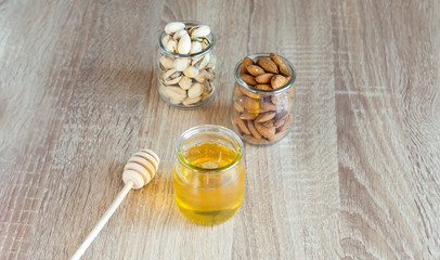 Honey in glass jar with wooden honey dipper and almonds, peanuts on light wooden table background. 