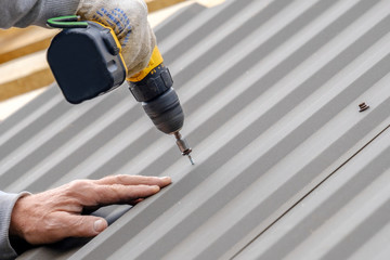 Men's hands in work gloves with a yellow screwdriver screw the roofing sheet to the roof of a country house. Cordless drill. The use of electrical engineering and technology.